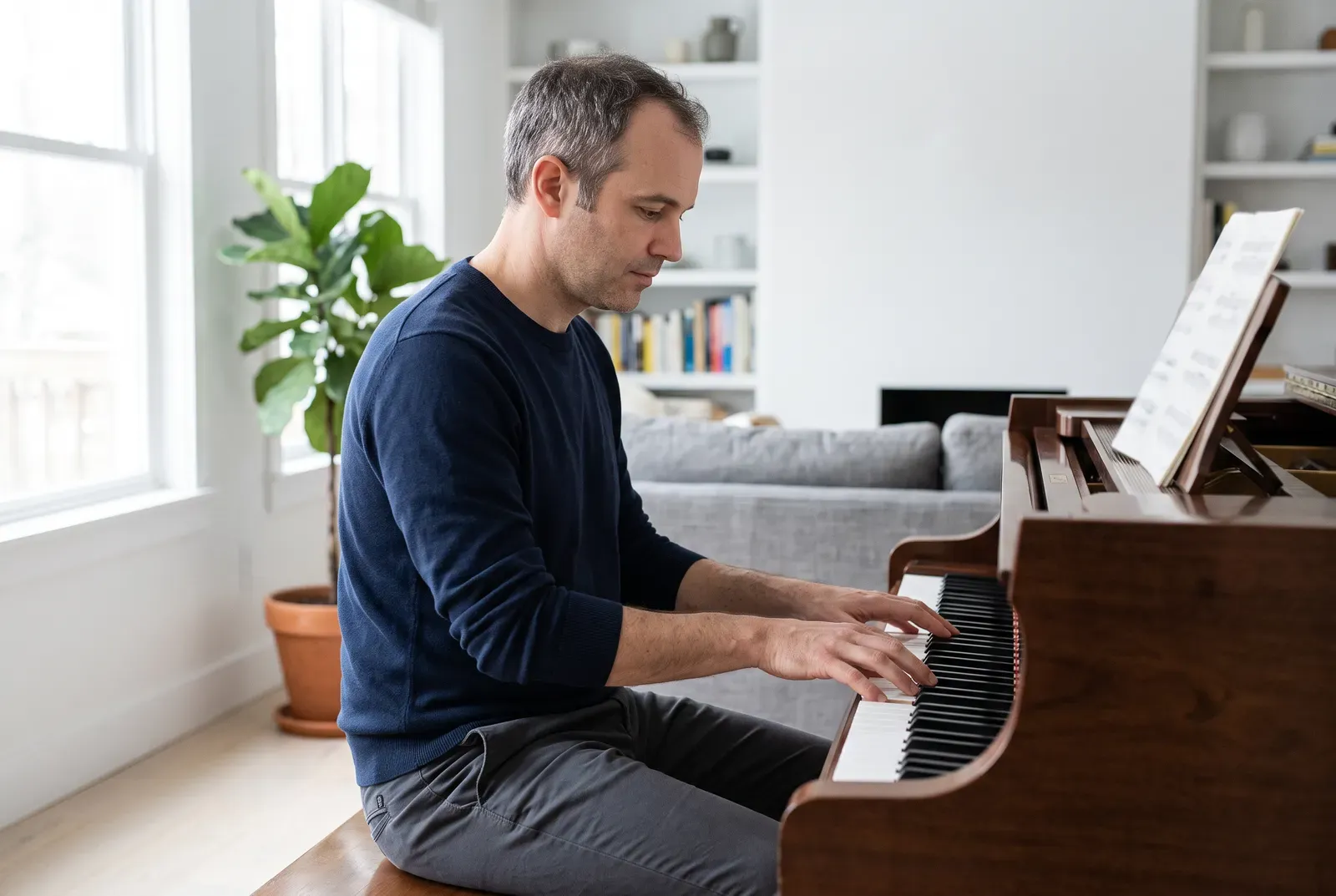 A man in his early forties sits at a piano in a bright living room, playing calmly and with focus.